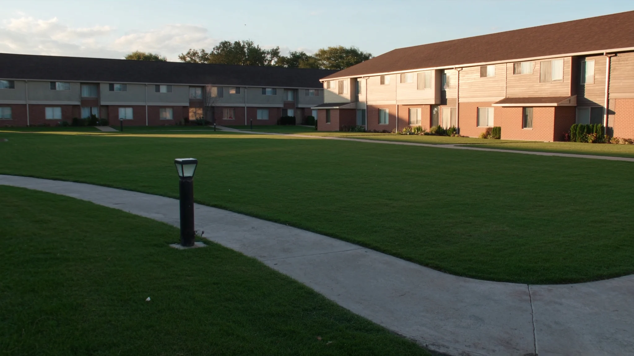 Open shared lawn between two rows of townhome buildings, with a concrete walkway curving across it in low late-afternoon light